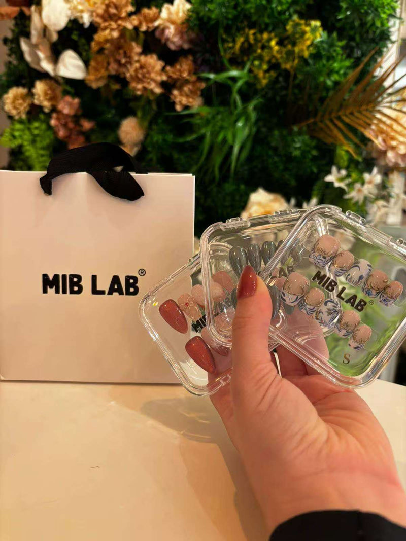Une main tient deux boîtes de faux ongles (press-on nails) MIB LAB, à côté d’un sac shopping MIB LAB, sur fond de mur végétal fleuri.