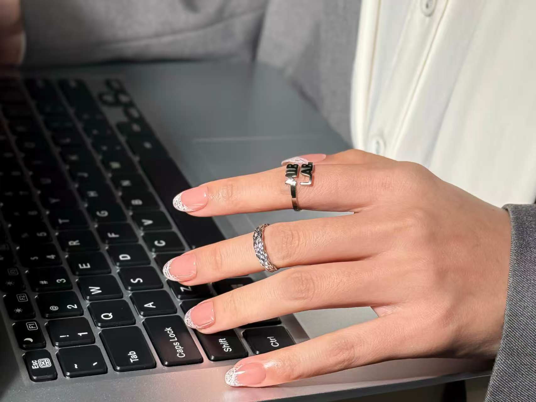Une main portant deux bagues tape sur le clavier d’un ordinateur portable, avec une manucure nude style french aux pointes blanches dentelées (ongles adhésifs).