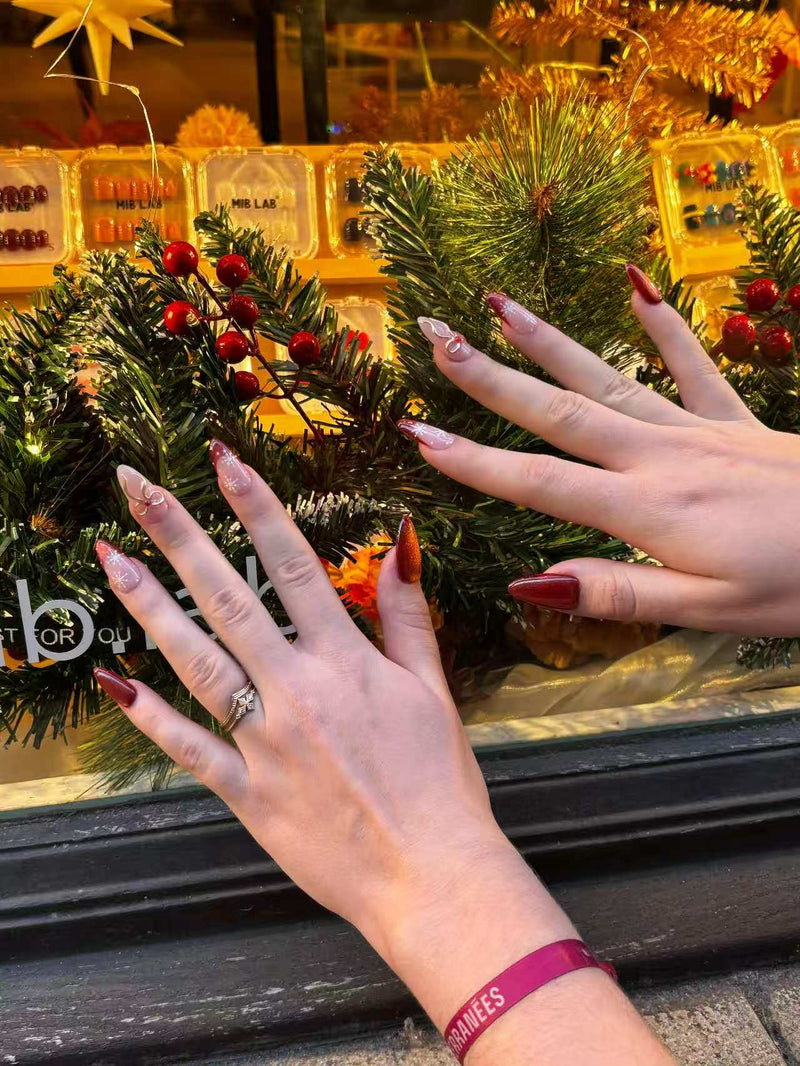 Deux mains présentent des press-on nails rouge brun avec flocons et nœuds, devant une décoration de Noël ; boîtes MIB LAB en arrière-plan.