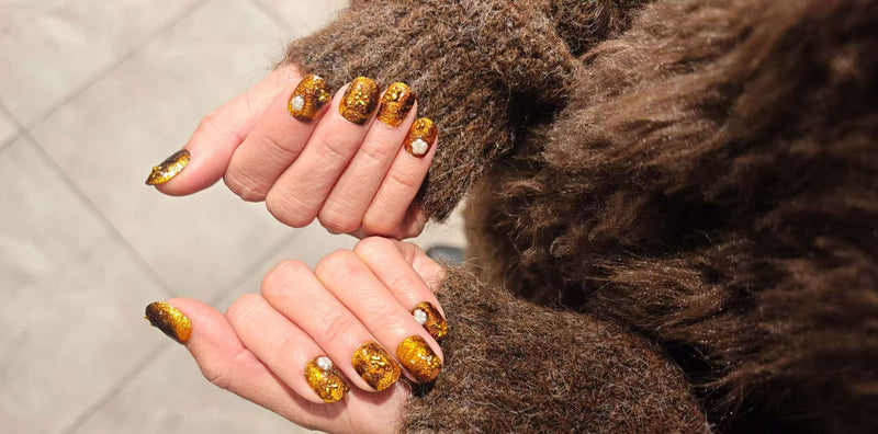 Gros plan sur deux mains avec des press-on nails dorés pailletés, décorés de petites perles, manches en fausse fourrure brune.