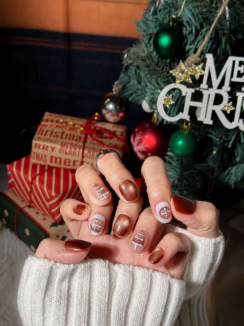 Gros plan des mains devant un sapin de Noël, avec des press-on nails marron et blancs aux motifs festifs (flocons/sapin).