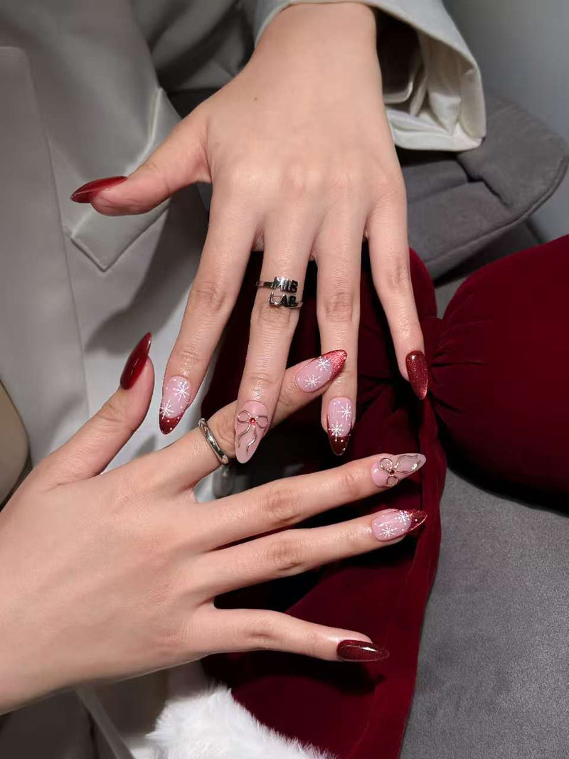 Deux mains présentent des press-on nails longs rouge bordeaux, avec détails flocons et nœuds sur base rose translucide.